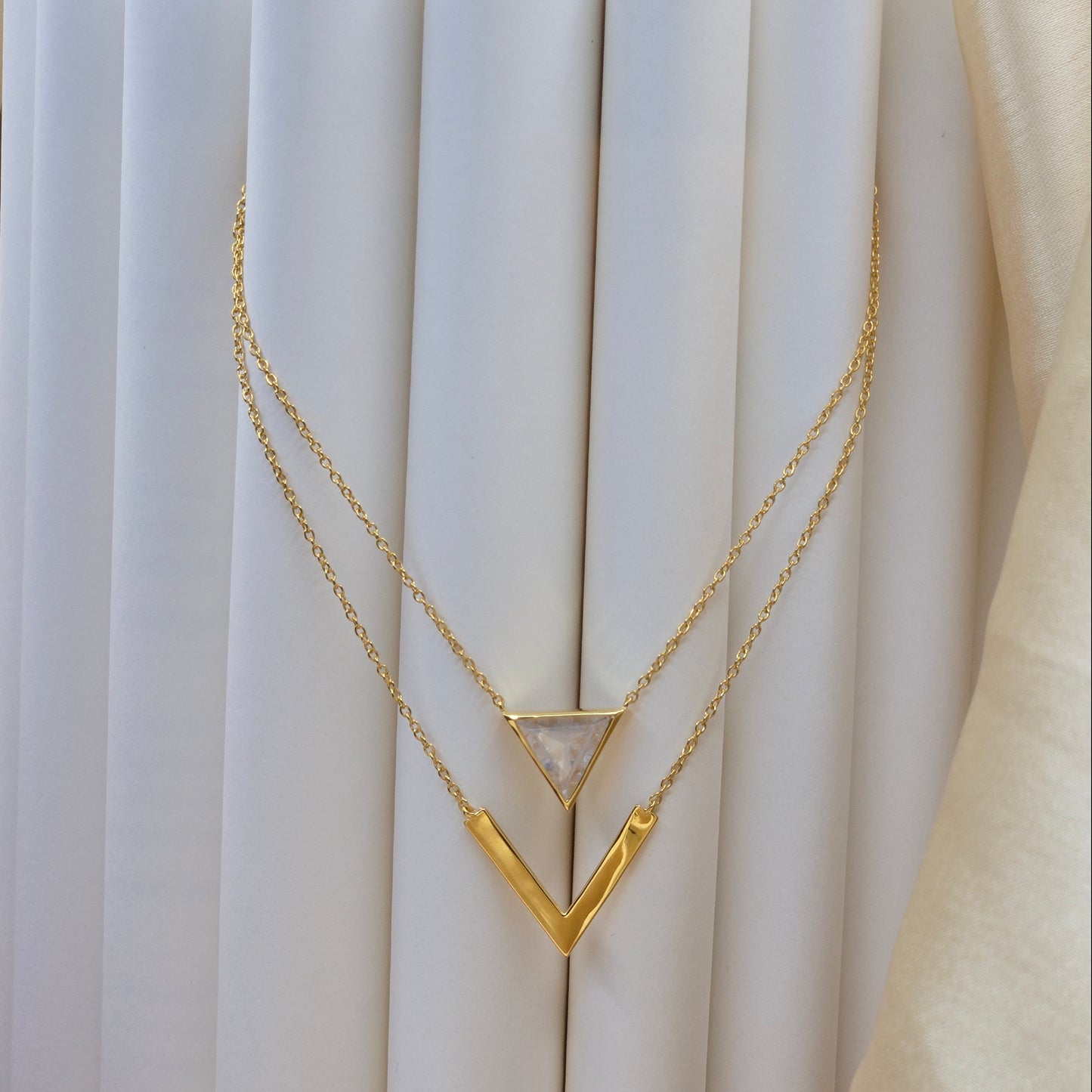 Layered gold-plated necklace featuring a clear triangular-cut stone pendant on the upper chain and a sleek V-shaped bar pendant on the lower chain, perfect for contemporary everyday styling.
