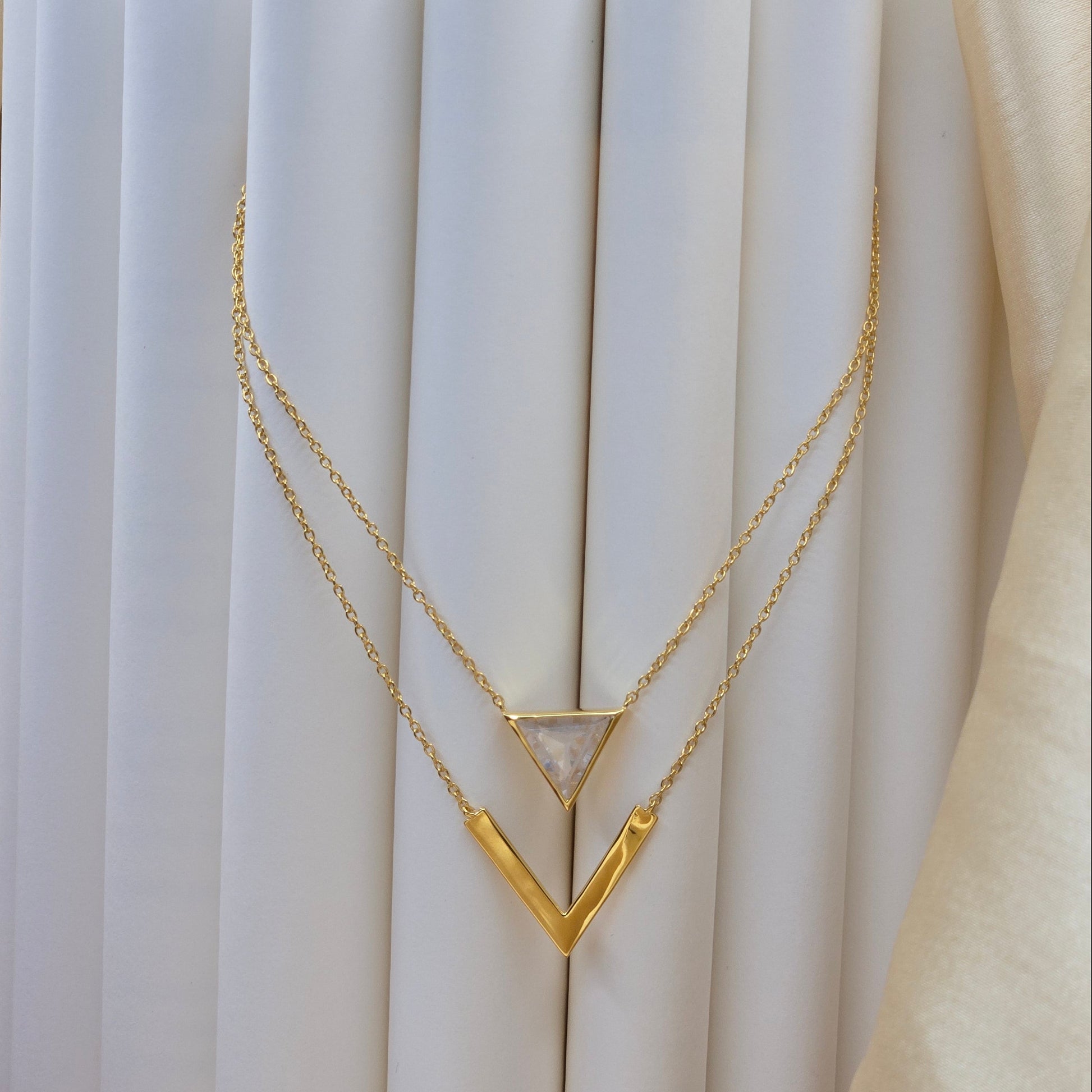Layered gold-plated necklace featuring a clear triangular-cut stone pendant on the upper chain and a sleek V-shaped bar pendant on the lower chain, perfect for contemporary everyday styling.