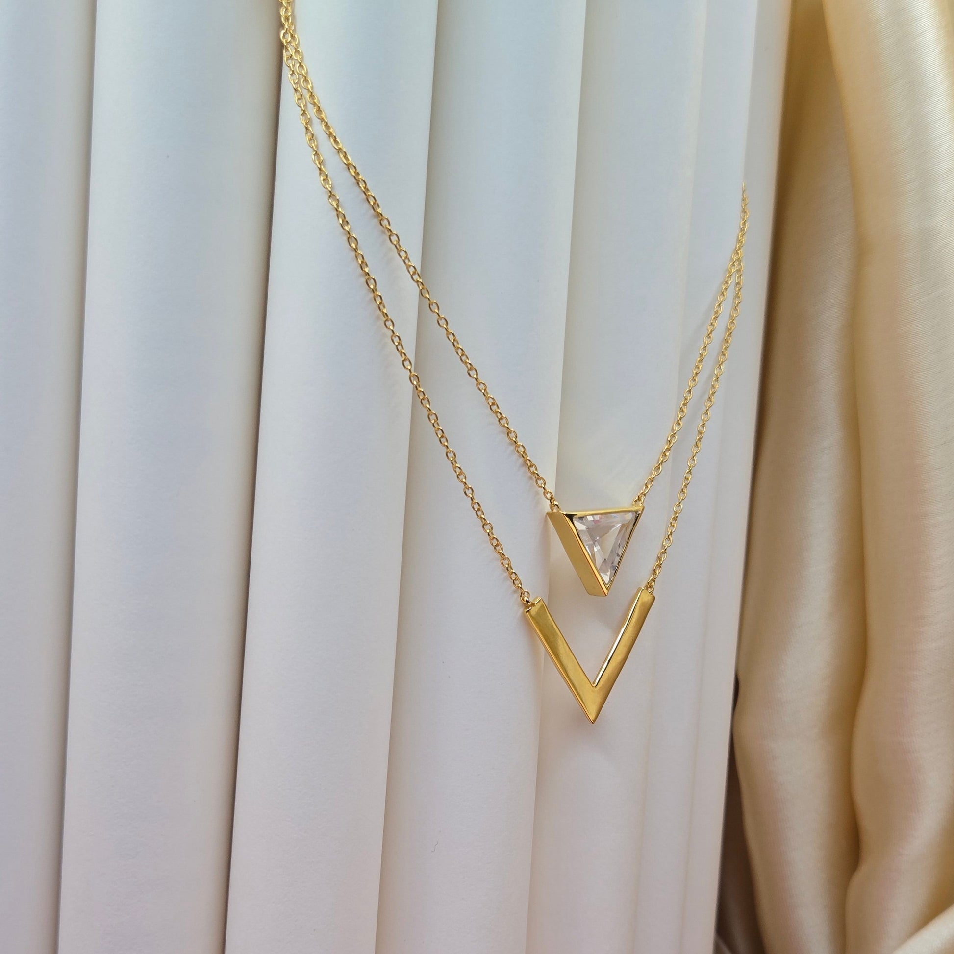 Layered gold-plated necklace featuring a clear triangular-cut stone pendant on the upper chain and a sleek V-shaped bar pendant on the lower chain, perfect for contemporary everyday styling.