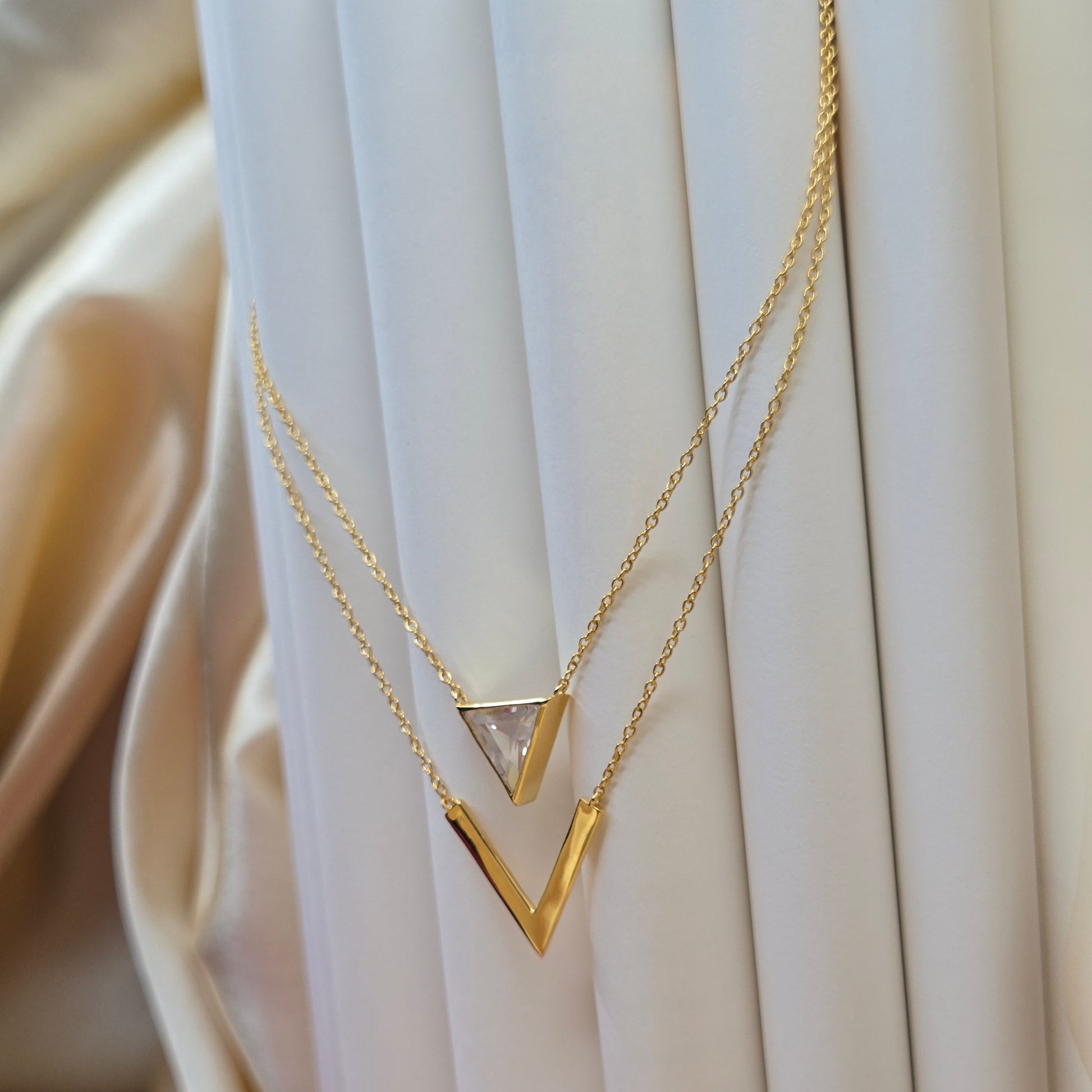 Layered gold-plated necklace featuring a clear triangular-cut stone pendant on the upper chain and a sleek V-shaped bar pendant on the lower chain, perfect for contemporary everyday styling.