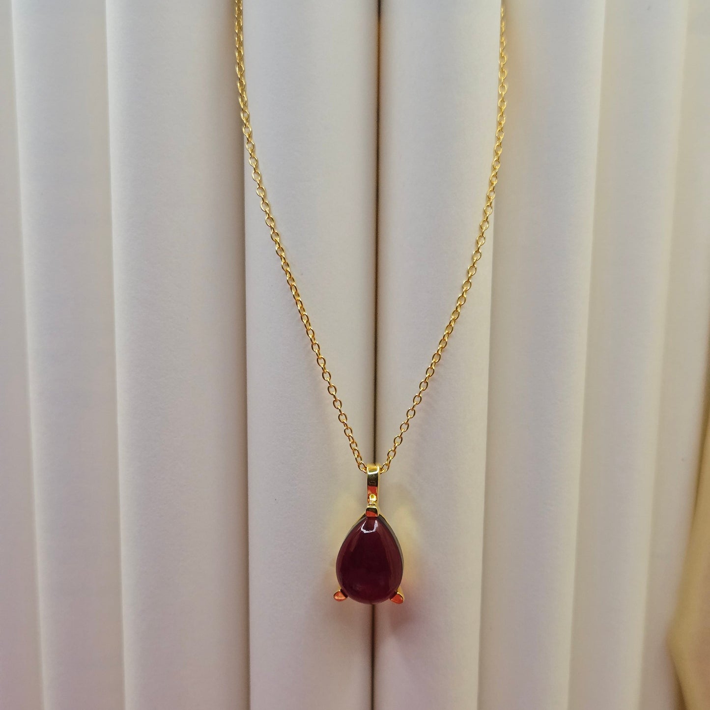 Gold-plated chain necklace featuring a smooth teardrop-shaped deep maroon gemstone pendant, styled against a soft neutral backdrop for an elegant and minimal look.