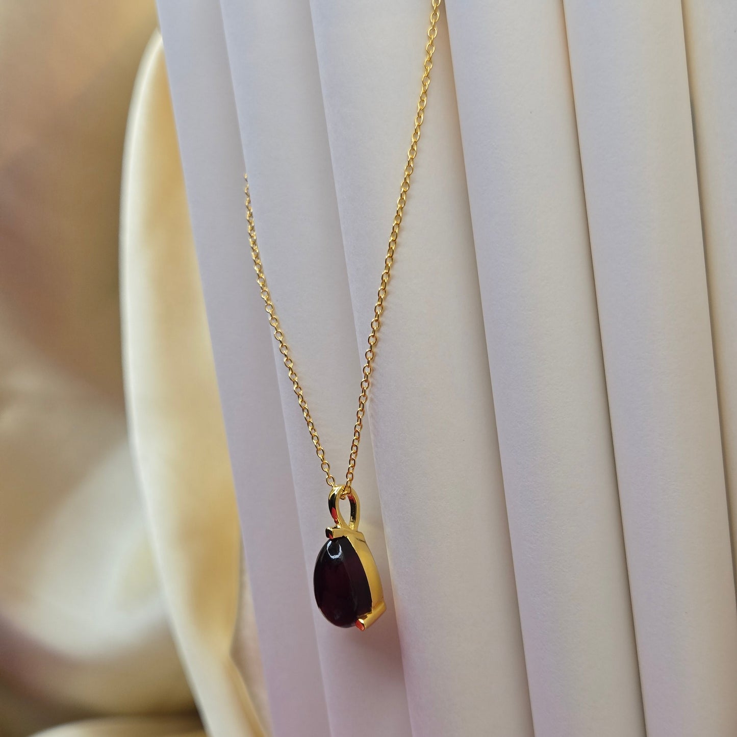 Gold-plated chain necklace featuring a smooth teardrop-shaped deep maroon gemstone pendant, styled against a soft neutral backdrop for an elegant and minimal look.
