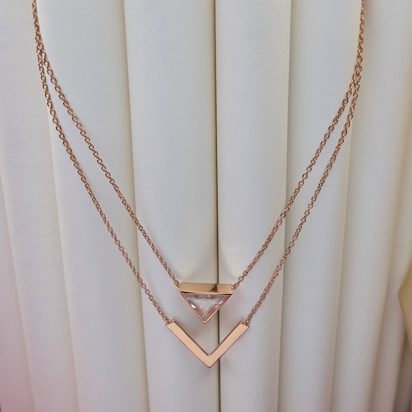 Layered rose-gold plated necklace featuring a clear triangular-cut stone pendant on the upper chain and a sleek V-shaped bar pendant on the lower chain, perfect for contemporary everyday styling.