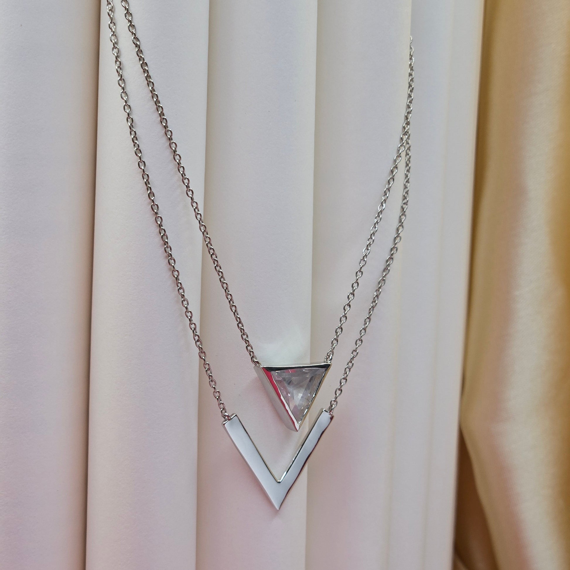 Layered rhodium plated necklace featuring a clear triangular-cut stone pendant on the upper chain and a sleek V-shaped bar pendant on the lower chain, perfect for contemporary everyday styling.