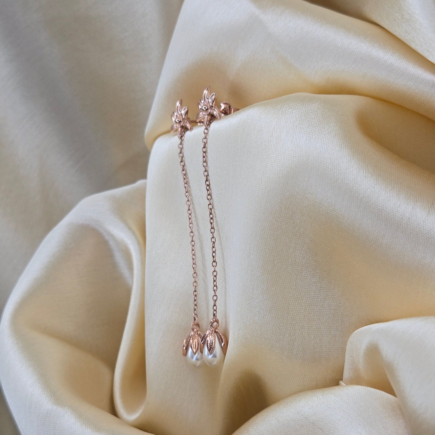 Lightweight rose-gold chain dangle earrings with pearl buds and intricate flower detailing, photographed on luxurious ivory satin for a refined and romantic aesthetic.