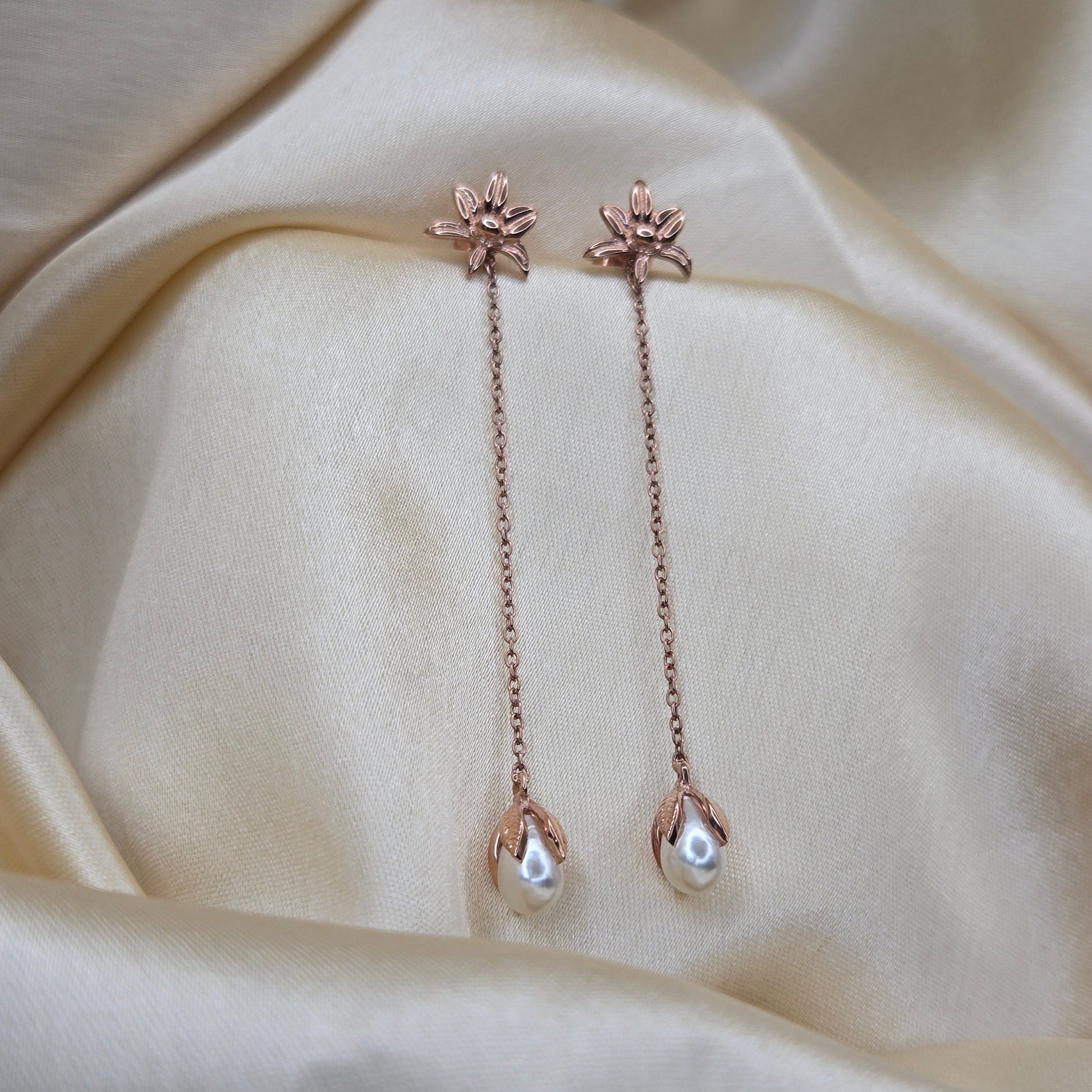 Lightweight rose-gold chain dangle earrings with pearl buds and intricate flower detailing, photographed on luxurious ivory satin for a refined and romantic aesthetic.